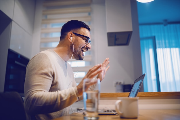 Het zijaanzicht van de mooie positieve mens kleedde toevallige zitting bij eettafel in keuken en hebbend videogesprek over laptop met zijn vriendin. Op tafel naast laptop staan glas water en koffie.