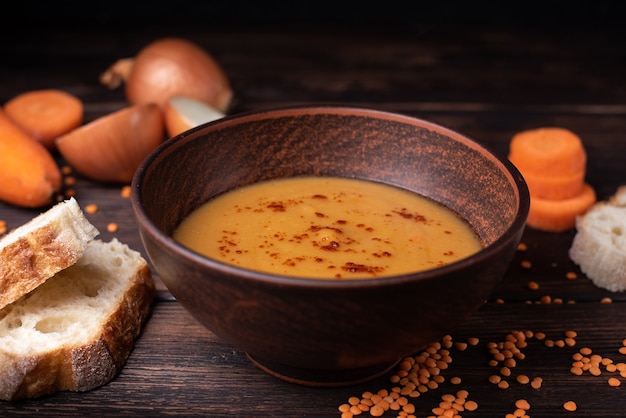 Hete heerlijke vegetarische rode linzensoep met ingrediënten: wortel, ui en brood op een donkere houten tafel, close-up.