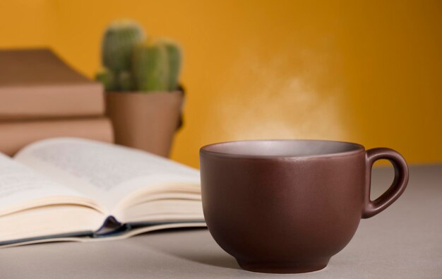 Hete stomende bruine koffie of theekop op bureau met open boek en cactus oranjegele achtergrond