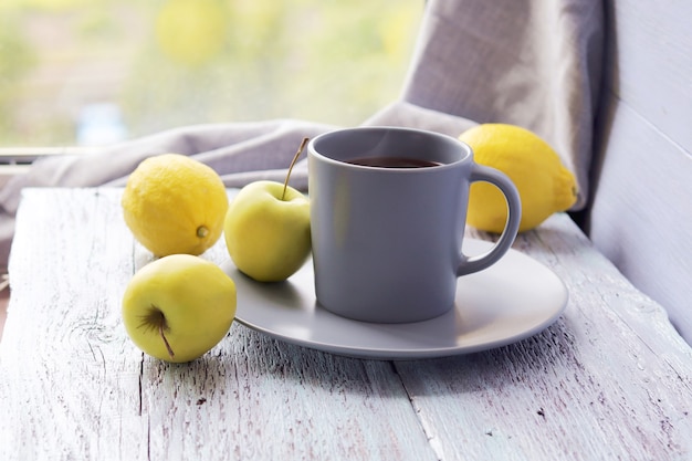 Hete thee in een grote kop, citroenen en appels op een houten tafel, gele en grijze kleur