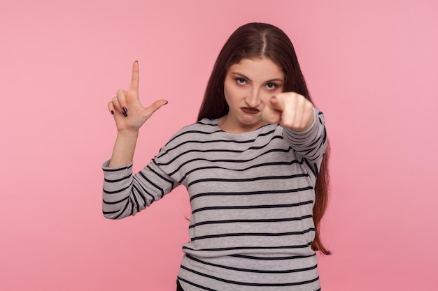 Hey je bent verloren portret van boze bazige vrouw in gestreept sweatshirt met L-teken verliezer gebaar en wijzend naar de camera die de schuld geeft van een mislukte nederlaag binnen studio opname geïsoleerd op roze achtergrond