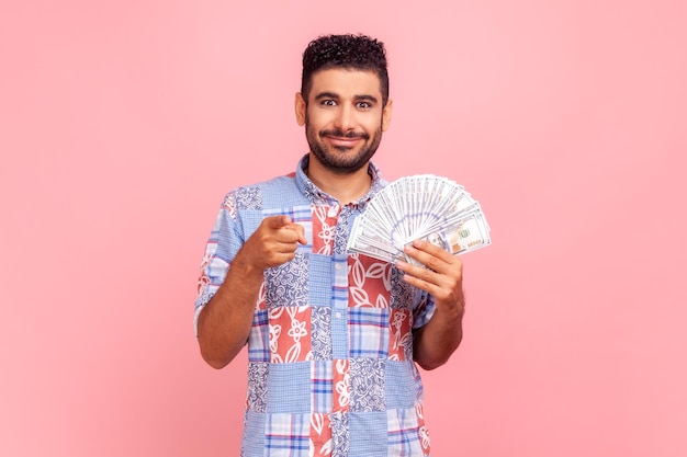 Hey je verdient geld Rijke man met blauwe casual stijl shirt wijzend naar de camera en met dollars kijken naar camera met brede glimlach Indoor studio shot geïsoleerd op roze achtergrond