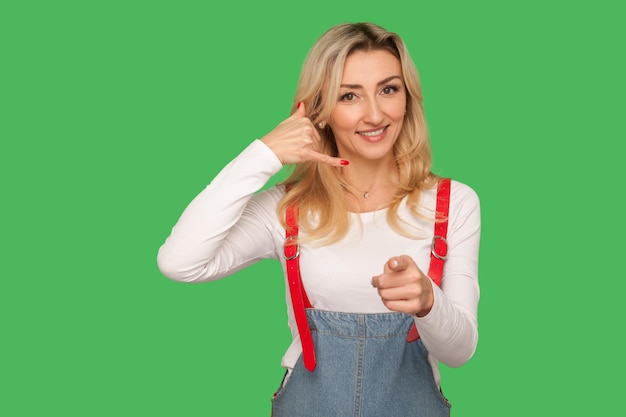 Hey jij, bel me terug, laten we telefonisch contact opnemen. portret van een speelse, gelukkige volwassen vrouw in een denim overall die een telefoongebaar maakt en naar de camera wijst. indoor studio-opname geïsoleerd op groene achtergrond