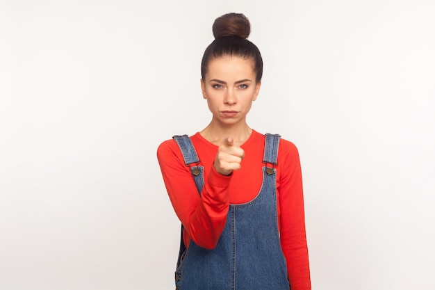 Hey jij Portret van een bazig ontevreden boos meisje met een haarbroodje in denim overalls wijzende vinger naar de camera die de schuld geeft en een kennisgeving geeft die streng is met schuldig studio-opname geïsoleerd