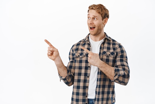 Hey look at this banner Smiling handsome redhead guy pointing and staring left aside at copyspace logo showing awesome deal in store demonstrate link on webpage standing over white background