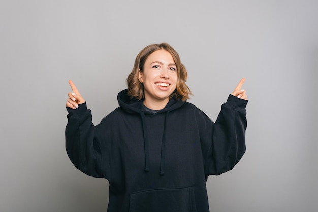 Hey look at this, young charming woman in black hoodie pointing up at copyspace over grey background