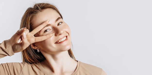 Hey smile you. Young beautiful girl smiles and shows peace victory gesture as fooling around on a white isolated background. A woman points to an idea, a place for advertising. Positive cute brunette