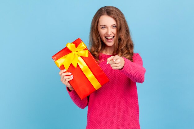 Hey you are winner Happy optimistic woman wearing pink pullover holding present box and pointing finger to camera with excited expression Indoor studio shot isolated on blue background