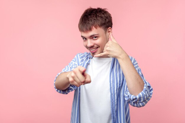 Hey you, call me back. portrait of positive brown-haired man with small beard and mustache in casual shirt making phone gesture and pointing at you. indoor studio shot isolated on pink background