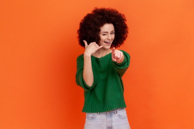 Hey you, call me. Woman with Afro hairstyle standing with telephone hand gesture and smiling to camera, flirting offering to contact by phone. Indoor studio shot isolated on orange background.