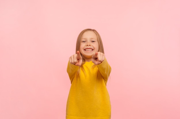 Hey you Cute happy little girl pointing to camera and smiling sincerely indicating lucky one choosing goods in child store meaning I need this indoor studio shot isolated on pink background