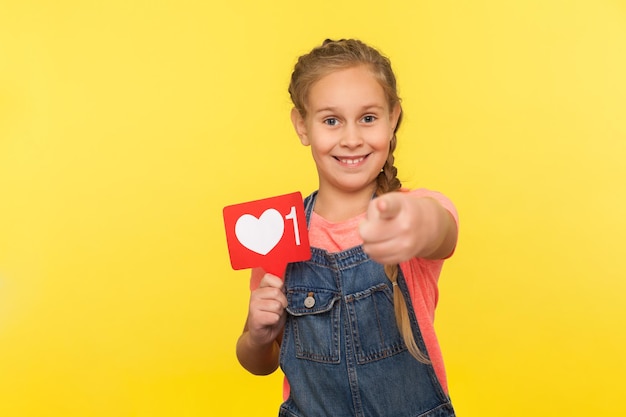 Hey you, like and comment child blog. Portrait of happy little girl in denim overalls holding social network heart button, follower notification symbol, popular internet content. studio shot isolated