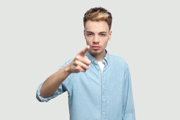 Hey you. Portrait of serious bossy handsome young man in light blue shirt standing, accusing and looking at camera with serious face. indoor studio shot on grey background copy space.