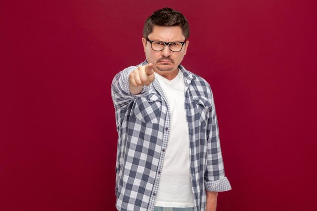 Hey you. Portrait of serious handsome middle aged business man in casual checkered shirt and eyeglasses standing, looking and pointing at camera. indoor studio shot, isolated on dark red background.