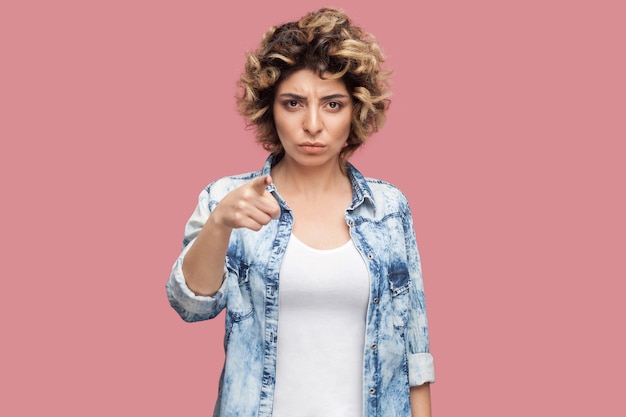 Hey you. Portrait of serious young woman with curly hairstyle in casual blue shirt standing, looking and pointing at camera with serious face. indoor studio shot, isolated on pink background.