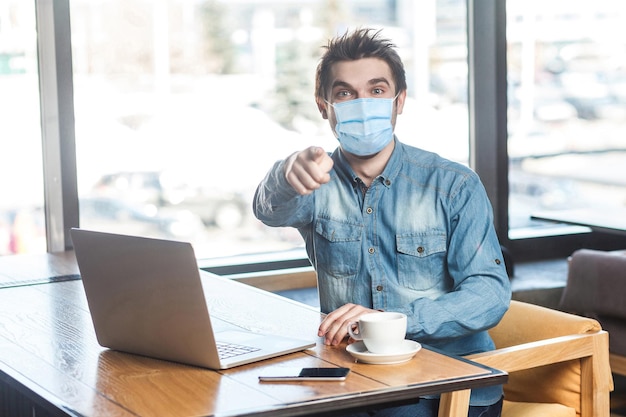 Hey you! Portrait of young man with surgical medical mask in blue shirt sitting and working on laptop and pointing finger to you, looking at camera. indoor working and health care concept.