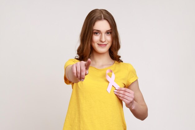 Hey you, take care of your health. Portrait of pretty optimistic teen girl in yellow T-shirt holding pink ribbon, symbol of breast cancer awareness. Indoor studio shot isolated on gray background.