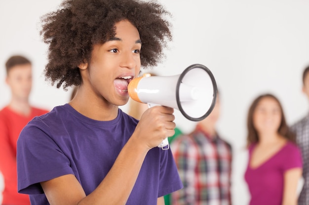 Hey! Young African teenager shouting at megaphone and looking away while his friends standing on background