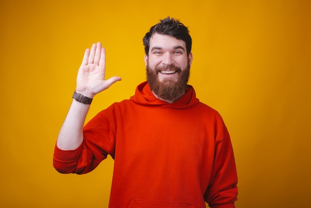 Hi friends, smiling man with beard in red blouse making Hello gesture