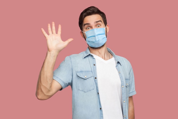 Ciao piacere di conoscerti. ritratto di giovane sorpreso con maschera medica chirurgica in camicia blu in piedi e guardando la telecamera con sorriso e saluto. foto in studio al coperto, isolata su sfondo rosa