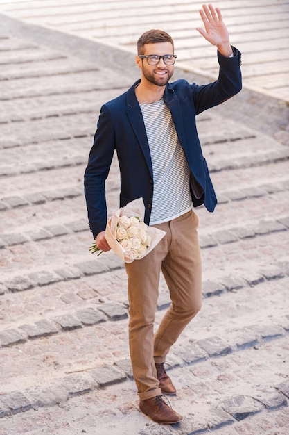 Всем привет! Веселый молодой человек в полный рост в элегантной повседневной одежде держит букет цветов и машет кому-то рукой во время прогулки по улице