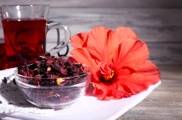 Hibiscus tea in glass teapot and flower on color napkin on wooden background