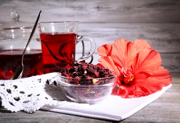 Hibiscus tea in glass teapot and flower on color napkin on wooden background