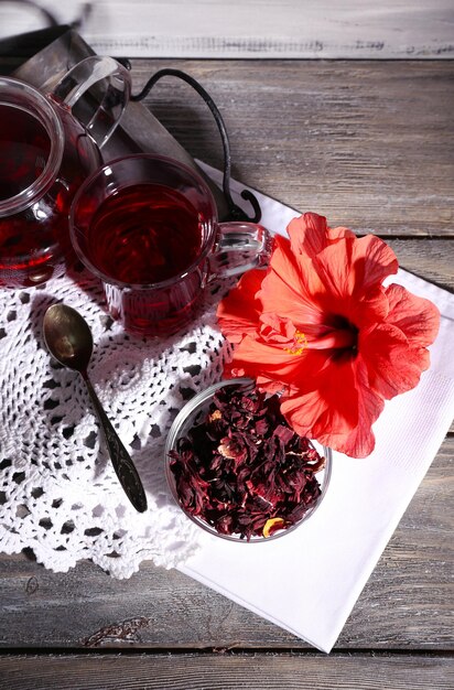Hibiscus tea in glass teapot and flower on color napkin on wooden background