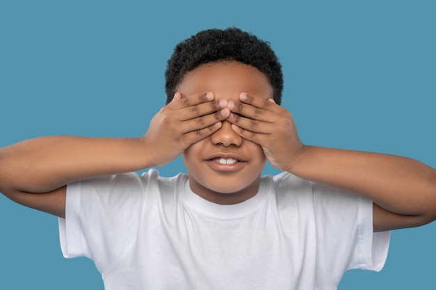 Hide and seek. african american boy of primary school age
standing with hands over his eyes on light blue background