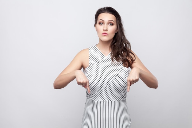 Hier en rechts weten. Portret van serieuze mooie jonge brunette vrouw met gestreepte jurk permanent en kijken naar camera met bazig gezicht. indoor studio opname, geïsoleerd op een grijze achtergrond.