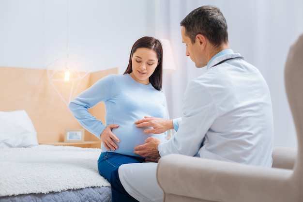 Hier is het. attente toekomstige mama die een glimlach op haar gezicht houdt en naar beneden kijkt terwijl ze op het bed zit