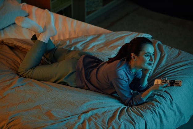 Взрослая женщина с высоким углом смотрит телевизор в темноте, лежа в постели дома.