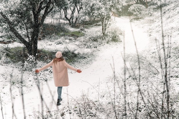 High angle back view of anonymous woman walking with outstretched arms along snowy path and enjoying stroll in forest in winter