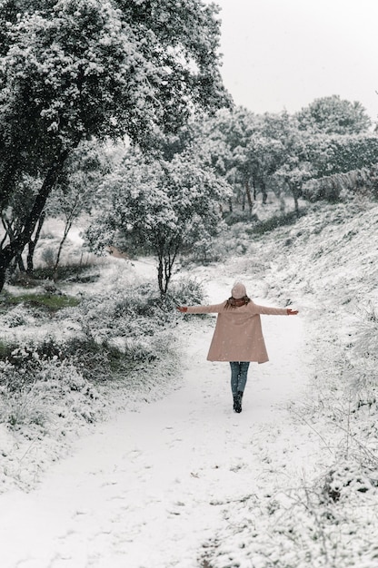 High angle back view of anonymous woman walking with outstretched arms along snowy path and enjoying stroll in forest in winter