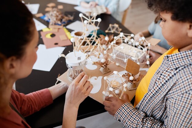 High angle closeup of African-American boy presenting school project to female teacher during art and craft lesson