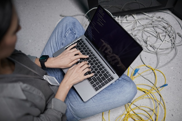 High angle closeup of female data engineer using laptop while sitting on floor in server room and setting up supercomputer network, copy space