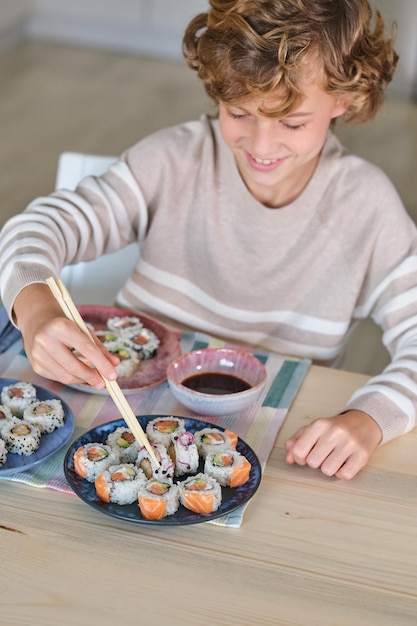 High angle of crop boy smiling and looking down while sitting at wooden table and eating sushi rolls with soy sauce using wooden chopsticks in kitchen at home