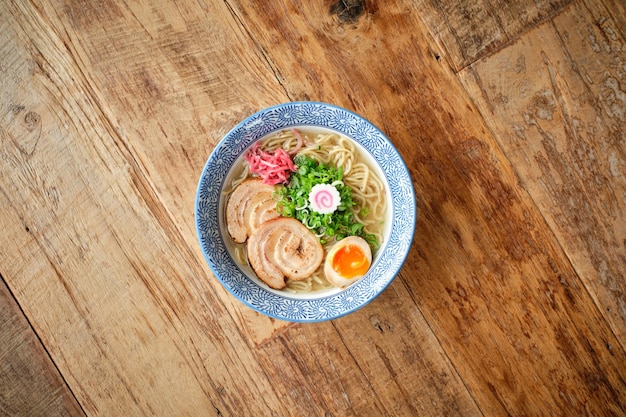 계란과 국수를 곁들인 맛있는 뜨거운 아시아 수프의 높은 각도는 나무 표면에 파란색 트레이서리가 있는 세라믹 접시에 제공됩니다.