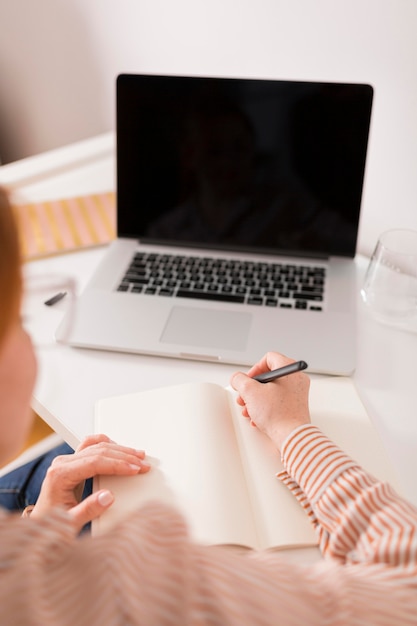 High angle of female teacher using laptop during online class and writing in agenda