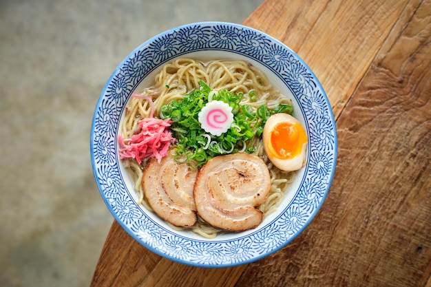 High angle of fresh cooked Japanese ramen with egg and sliced pork served in ceramic bowl placed on edge of round shaped wooden table