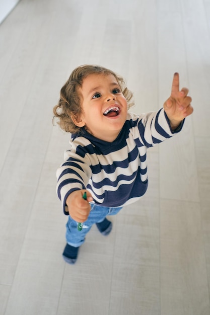 High angle full body of adorable excited toddler with curly hair in casual clothes standing on floor and pointing up while holding colorful marker