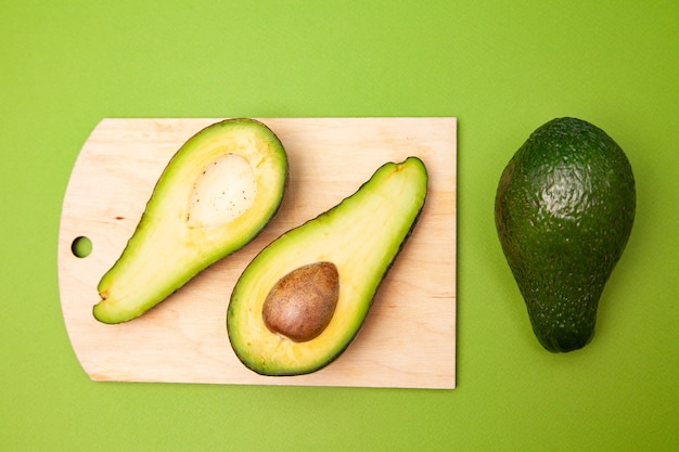 High angle of halved avocado with seed placed on wooden cutting board near whole fruit on green background