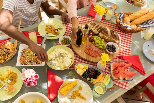 写真 太陽の下、外のパティオで家族で食事中にテーブルに座って、食べ物を出したり食べたりする混血カップルの高角中央断面図