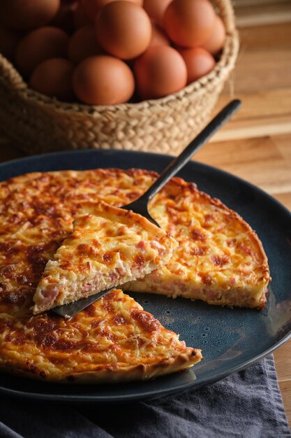 High angle of plate with tasty cheese and bacon quiche placed on table near wicker basket with fresh eggs