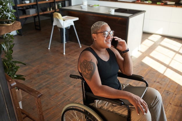 High angle portrait of contemporary tattooed woman with disability speaking by smartphone while working from home, copy space