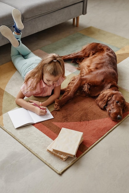 犬と一緒に床に横たわっている間宿題をしているかわいいブロンドの女の子のハイアングルの肖像画