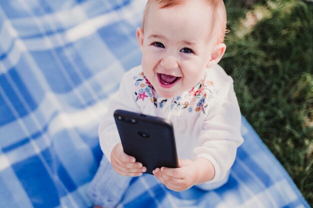 Foto ritratto ad alto angolo di una bambina sorridente che tiene in mano il telefono su una coperta da picnic