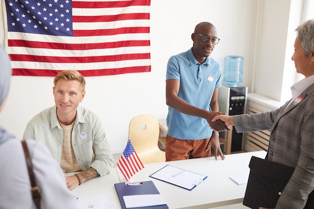 High angle portrait of two polling station workers shaking hands with people while registering voters on election day, copy space