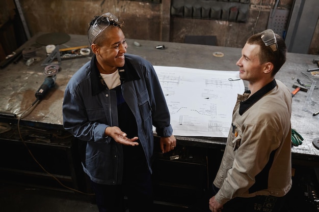 High angle portrait of two smiling factory workers discussing project while standing in workshop