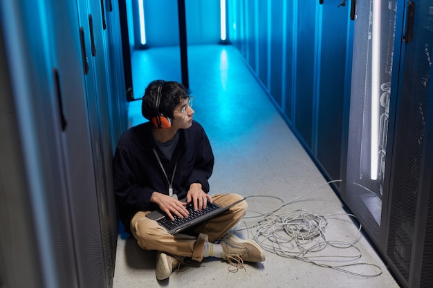 High angle portrait of young Asian man sitting on floor in server room lit by blue light while setting up supercomputer network, copy space